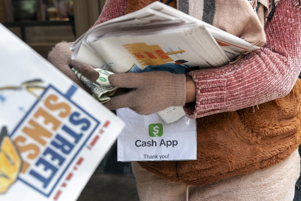 A vendor selling Street Sense, a local paper that covers issues related to the homeless and employs unhoused individuals as its vendors, takes a donation for a paper while wearing a sign saying she can accept donations from cashless apps like Venmo and CashApp, Wednesday, Dec. 6, 2023, in Washington. The transition to a cashless society has impacted street-level charitable giving – from individual donations to panhandlers to the Salvation Army donation kettles. (AP Photo/Jacquelyn Martin)