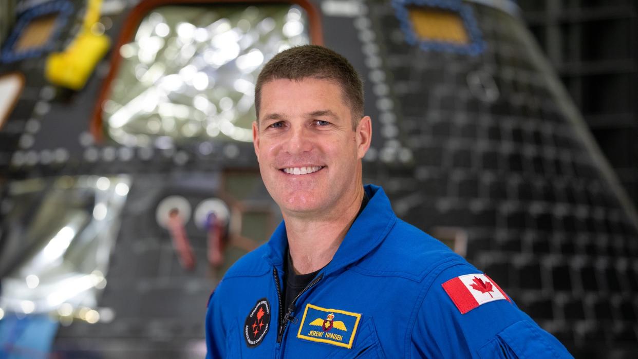  jeremy hansen smiling and standing in a flight suit. the canadian flag is visible on his left shoulder. behind him is a blurry, cone-shaped image of a spacecraft and you can see silver foil covering the window shining 