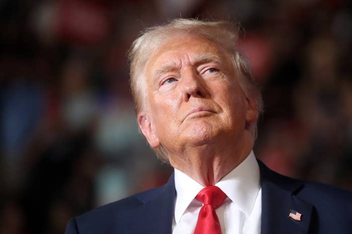 Donald Trump in a red suit and tie, looking slightly upward, in a public setting