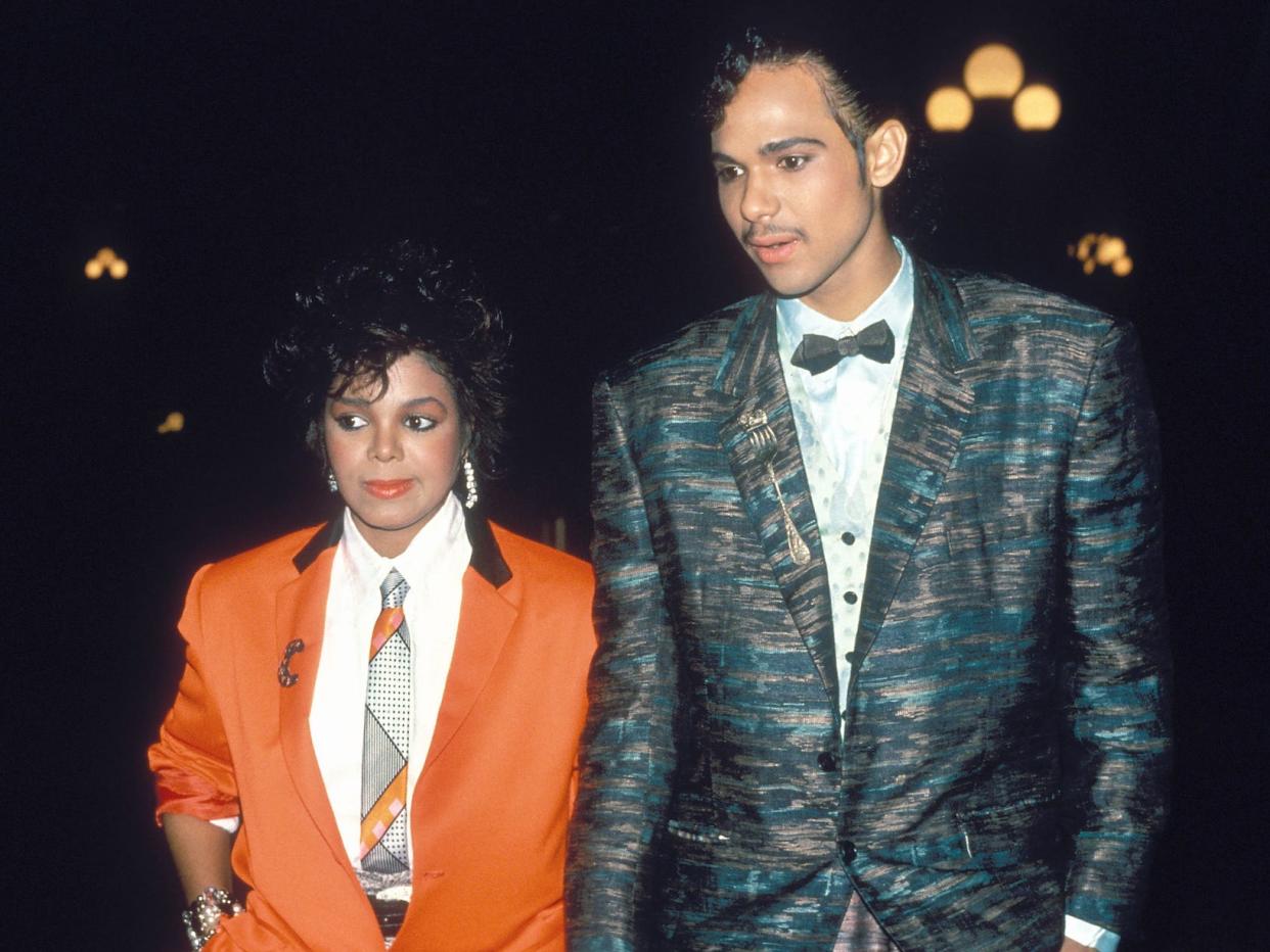 A young Black woman (Janet Jackson) and a young Black man (James DeBarge) photographed together in 1984. Jackson wears a red suit jacket with a tie and white shirt, while DeBarge has a blue patterned suit on with a light blue shirt and black bow tie.