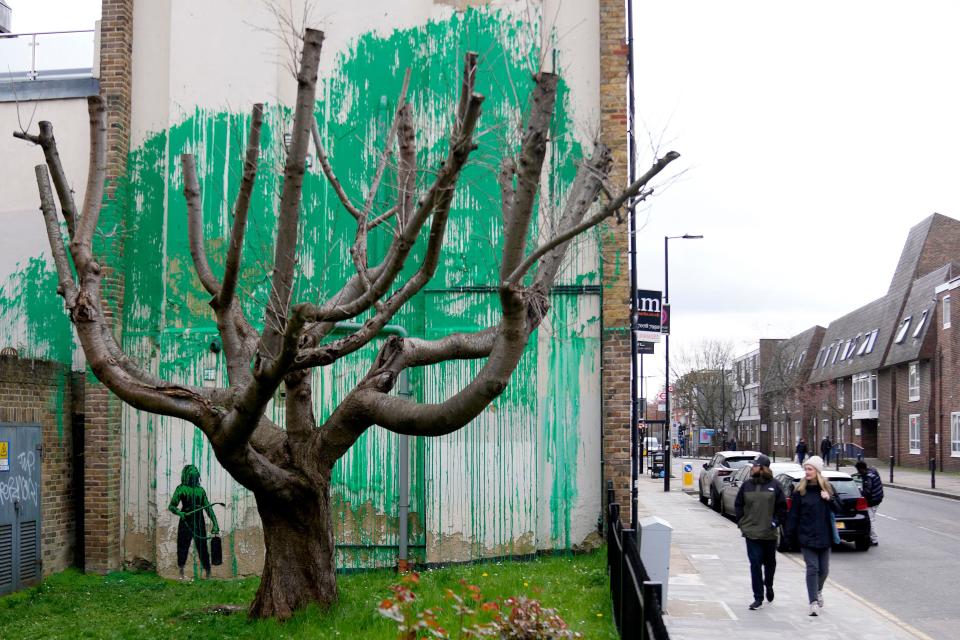 People look at a new Banksy painting on a wall in London, Monday, March 18, 2024. A new Banksy mural drew crowds to a London street on Monday, even before the elusive graffiti artist confirmed that the work was his. The artwork in the Finsbury Park neighborhood covers the wall of a four-story building and shows a small figure holding a pressure hose beside a real tree. Green paint has been sprayed across the wall, replicating the absent leaves of the tree, which has been severely cropped. Banksy claimed the work by posting before and after photos of the location on his official Instagram accou