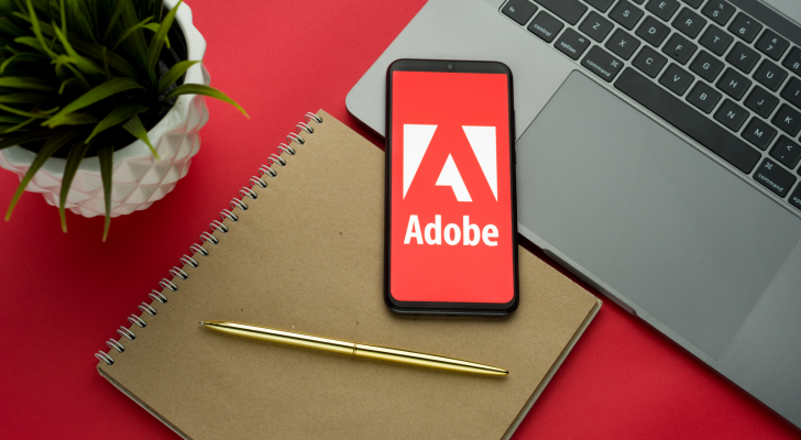 Adobe logo on the smartphone screen is placed on the Apple macbook keyboard on red desk background. ADBE stock.