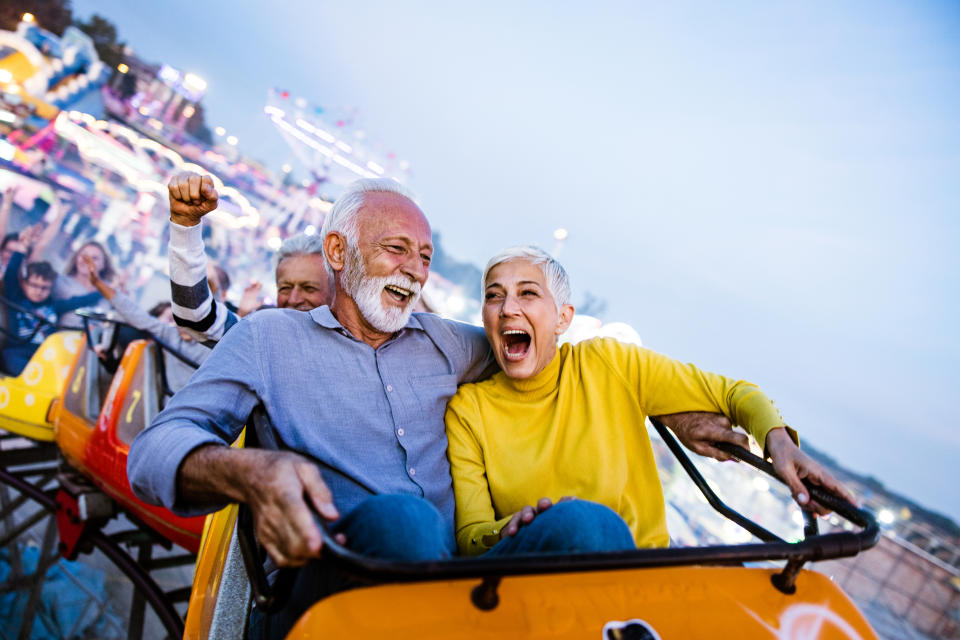 Elderly couple enjoys a rollercoaster ride, smiling and laughing, with an amusement park in the background