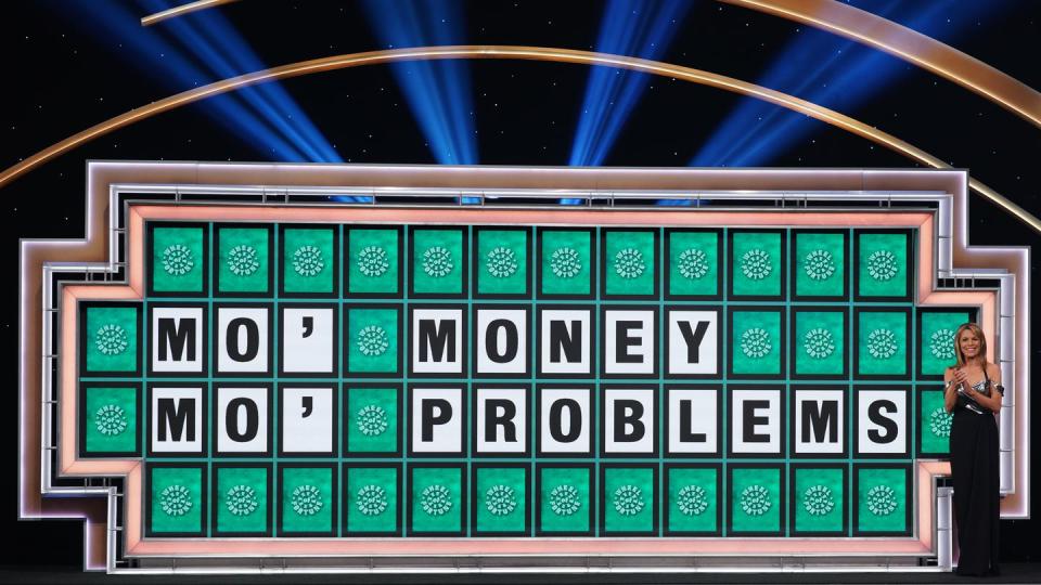 vanna white standing in front of a wheel of fortune puzzle that reads mo' money mo' problems