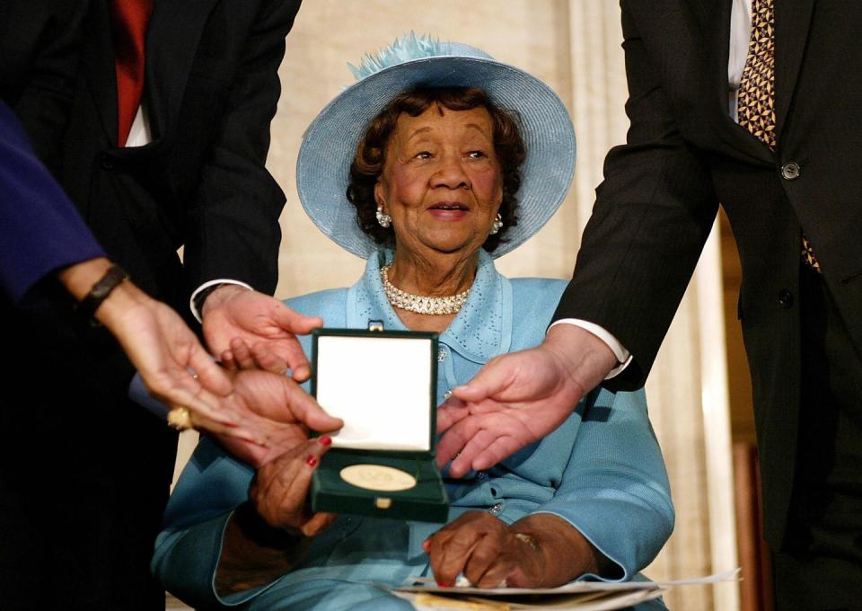 dorothy height sits and holds a gold medal in a small box along with several other hands, she wears a blue suit and matching hat with large earrings and a matching necklace