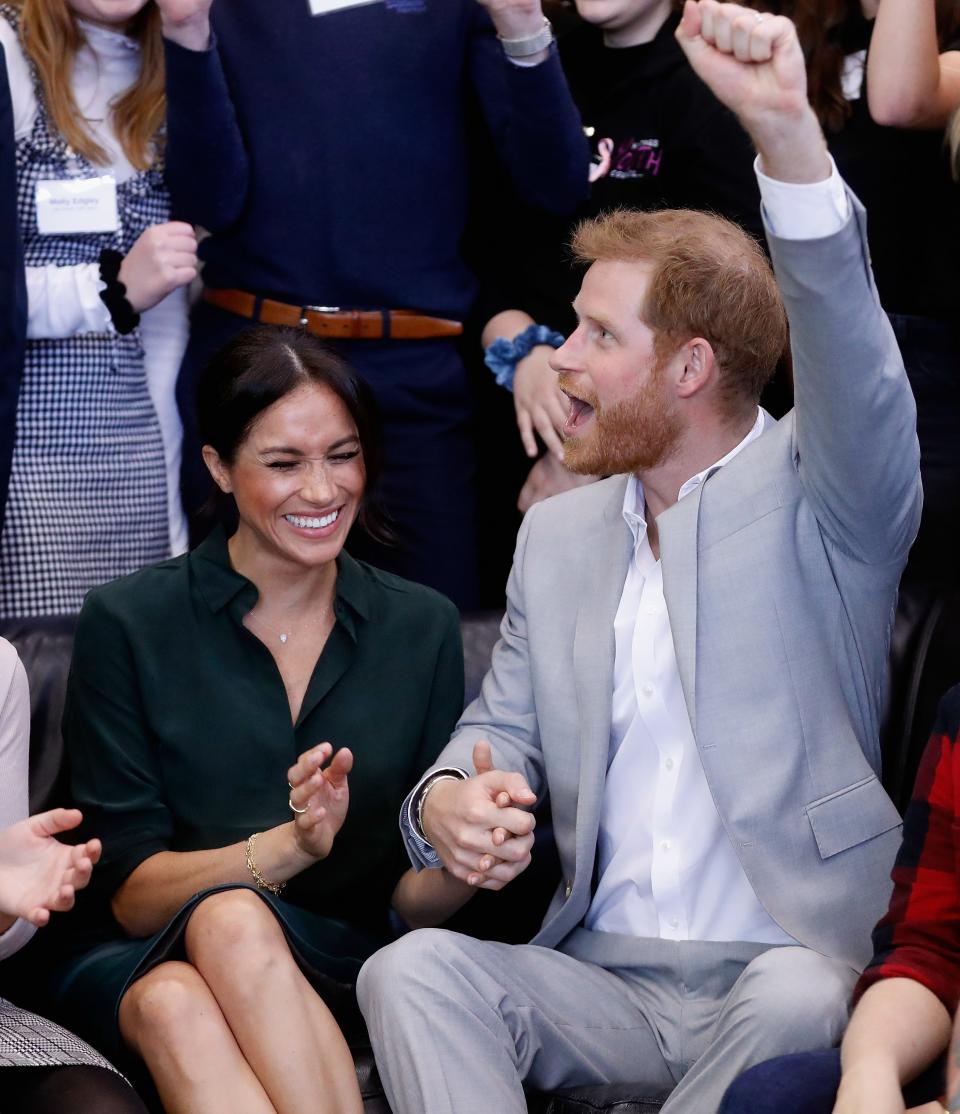 Meghan and Harry sitting and holding hands. Meghan smiles with her eyes closed and Harry cheers with his fist up in the air.