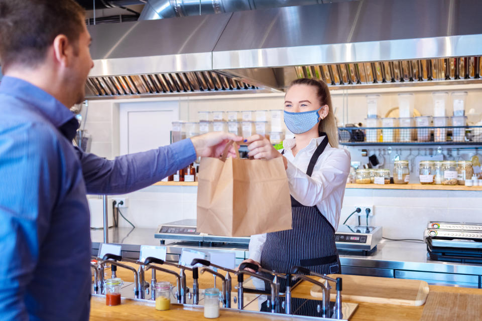 Giovane cameriera che indossa maschera protettiva e grembiule che serve i clienti al bancone in un piccolo ristorante - Concetto di piccola impresa e imprenditore con proprietaria di donna in un ristorante con consegna del servizio da asporto