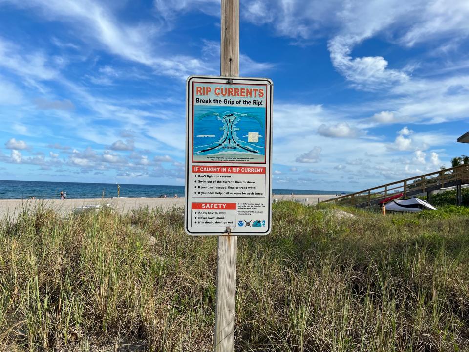 A beach sign in front of a grassy area and the sand warning of rip currents.