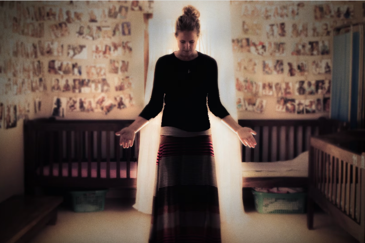 A photo shows a white woman standing in a room surrounded by small children's beds and out of focus photos. Her head is down, and her hands are out to each side.
