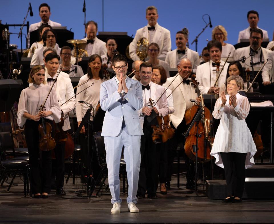 A man in a light blue suit stands onstage in front of standing orchestra members