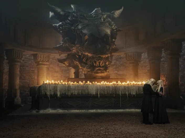 A wide shot of a room with a big dragon skull at its center, light by candles underneath. An old mad and his daughter stand close to each other in front of the skull.