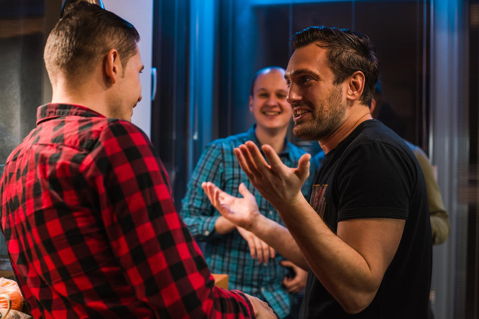 Three men, including one in a red plaid shirt, engaged in a lively conversation, standing together indoors