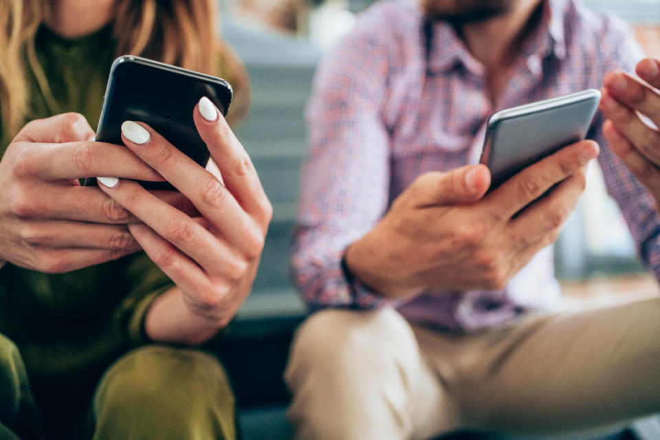 Man and woman sitting, holding smartphones, both looking at their screens. The woman's nails are painted white