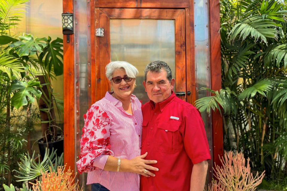 A woman and a man stand outside the door to a greenhouse in their yard