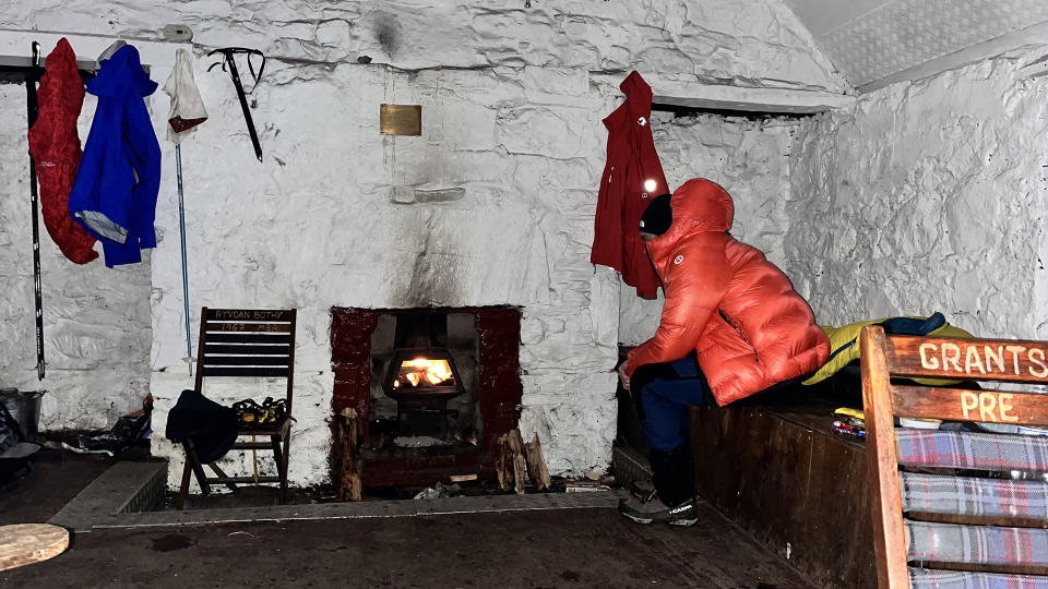 A man sits in a bothy wearing an orange The North Face Summit Series Pumori Down Parka