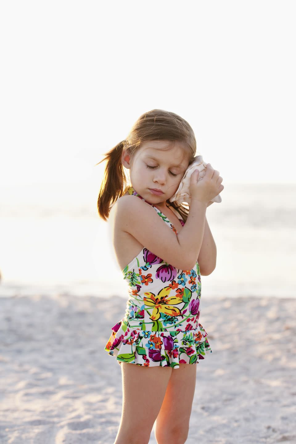 girl in floral print swimsuit listening to seashell at beach