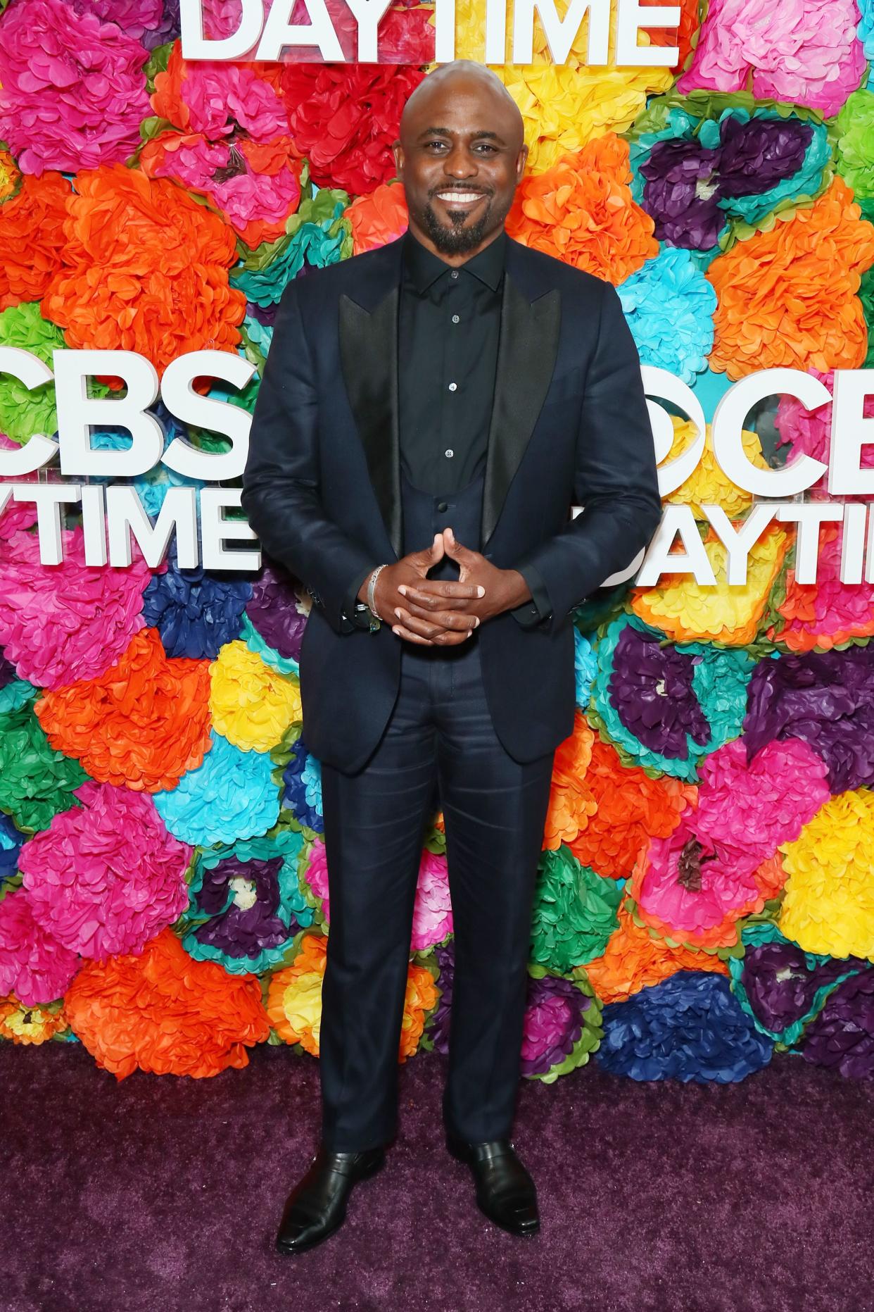 Wayne Brady wears a suit jacket and stands in front of a floral wall