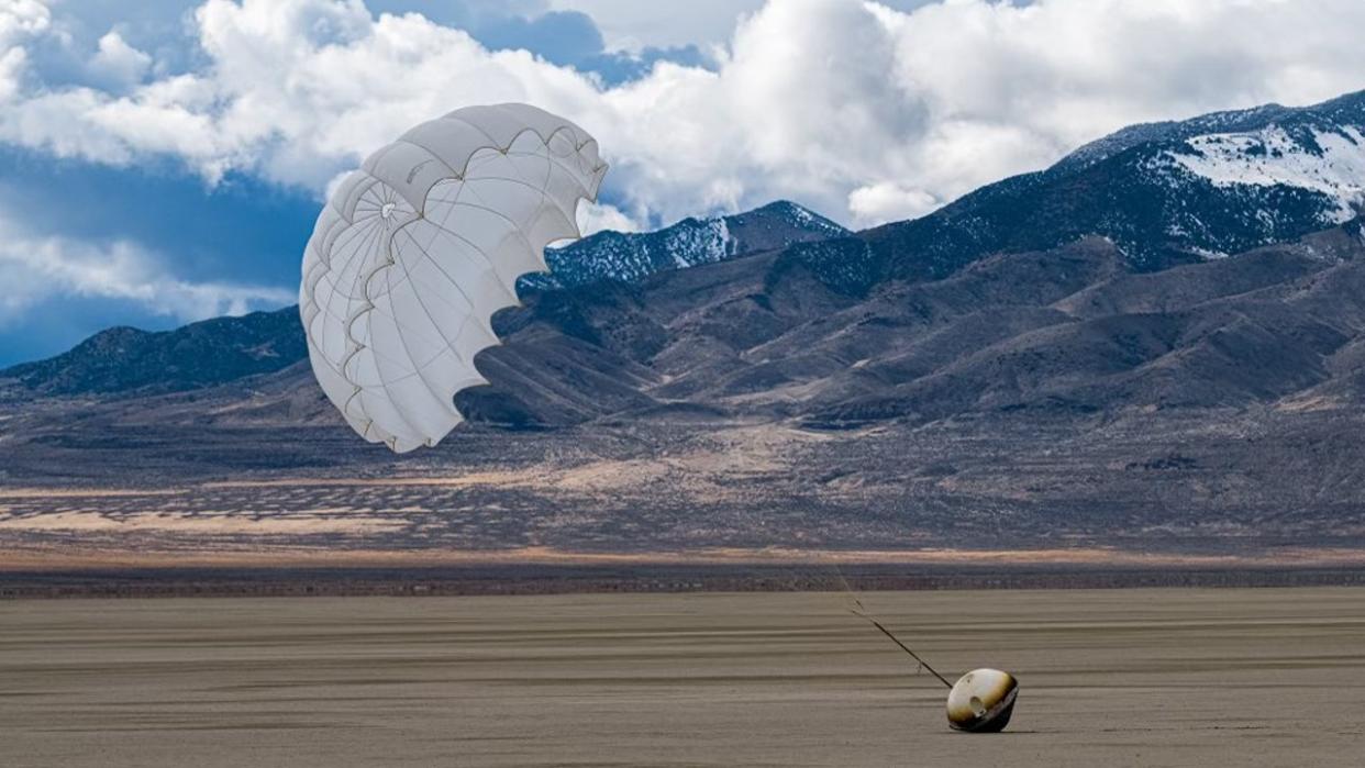  A cone-shaped capsule lies on the desert floor with a parachute billowing in the wind above it. 