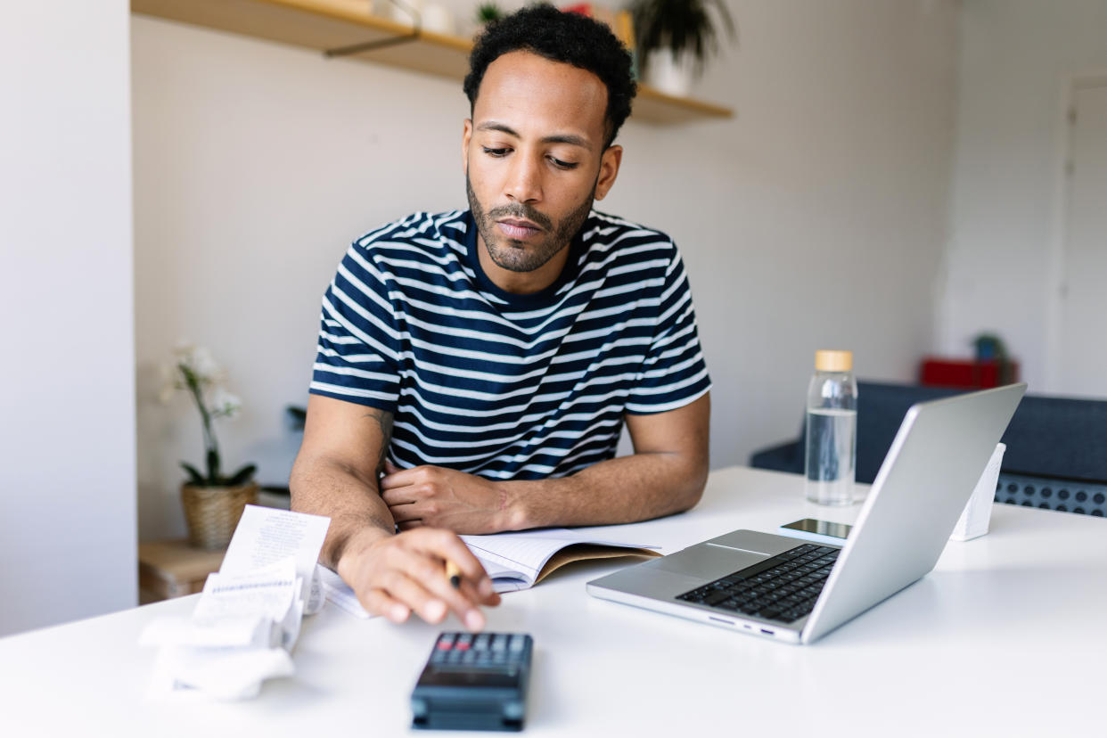 Young adult man with laptop checking bills, taxes, bank account balance and calculating expenses sitting at living room table.