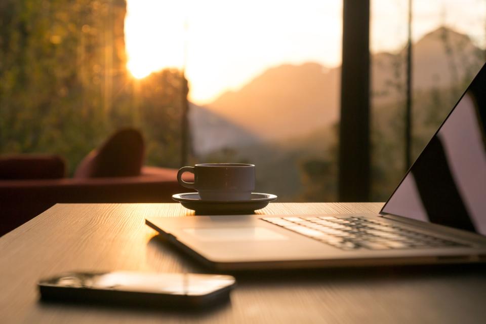 A laptop, smartphone, and cup of coffee sitting on a table. A window with sunshine shining through is in the background.