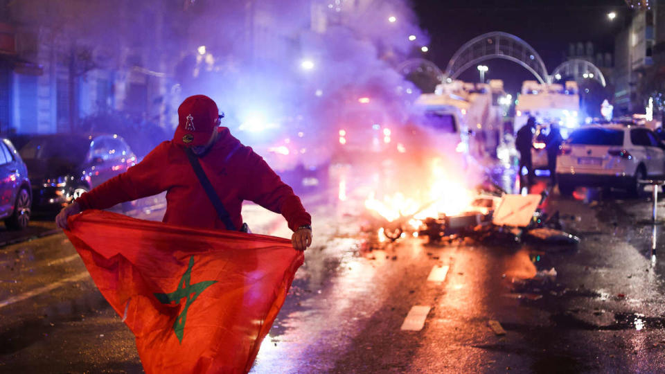 Un supporter avec un drapeau marocain alors que des scooters électriques brûlent dans son dos, en marge du match de la Coupe du monde 2022 entre la Belgique et le Maroc, à Bruxelles.