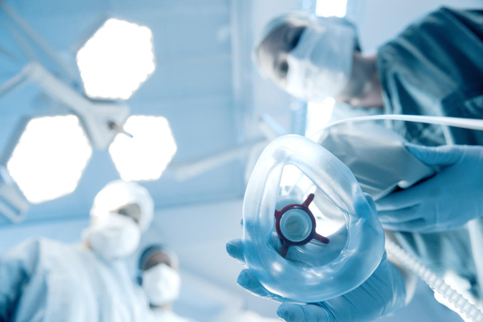 Three medical professionals, wearing surgical masks and scrubs, prepare to administer anesthesia through a mask, viewed from the patient's perspective