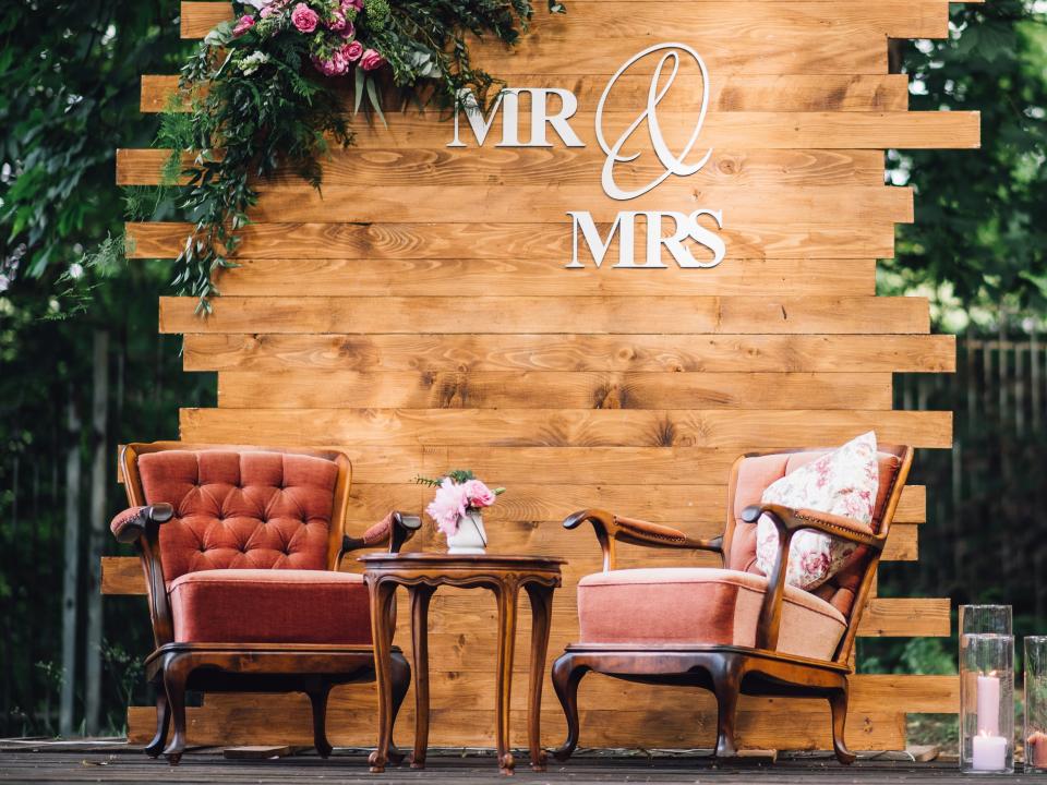 A wooden panel that says "Mr. & Mrs." and a floral arrangement on it with wooden chairs with cushions in front of it