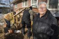 Elaine Fleming, of Cass Lake, Minn., stands outside of her home where she takes in stray and abandoned cats. In the background, friends Rick Haaland and Preston Lassman stuff straw under Elaine's front steps to provide shelter for the cats on Sunday, Nov. 21, 2021. Fleming and Haaland have been leading efforts among their fellow Ojibwe community members to care for animals, something centrally important to Native American spiritual beliefs and traditions. (AP Photo/Jack Rendulich)