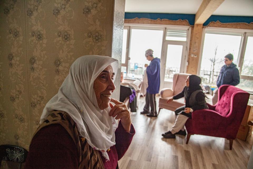 A woman in a white headscarf smiles near a wall; in the background are three people gathered near armchairs