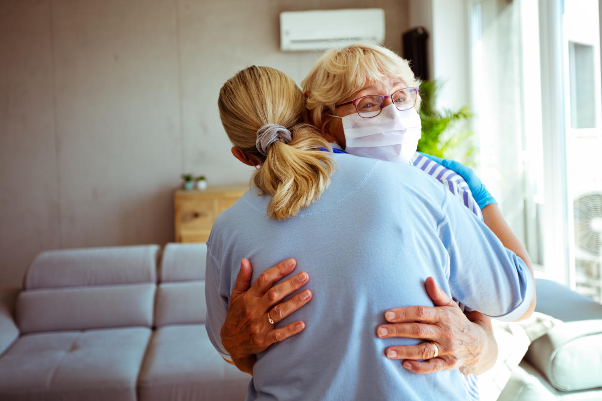 District nurse embracing worried with senior woman during home visit. They are standing in living room.