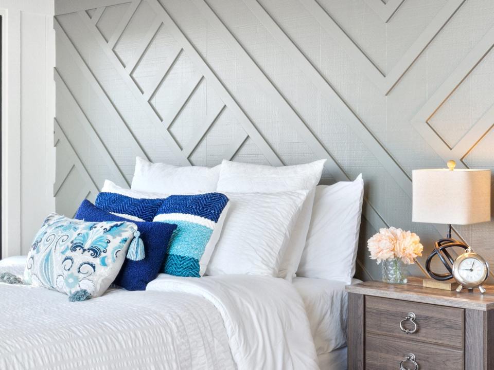A bed with white sheets and blue pillows and an end table with a lamp and a gray wall with diagonal paneling behind bed