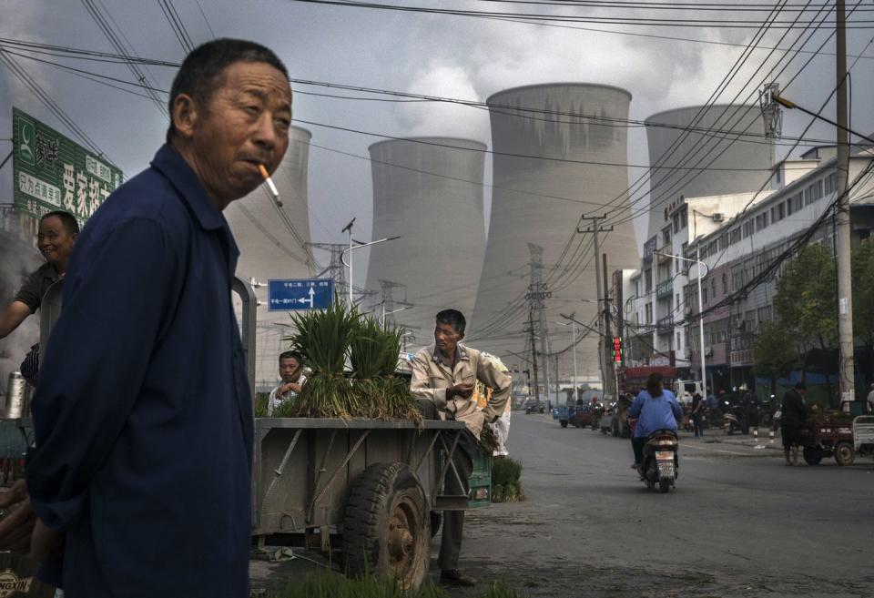 A man stands in a street market smoking, with cooling towers for a power plant behind him.
