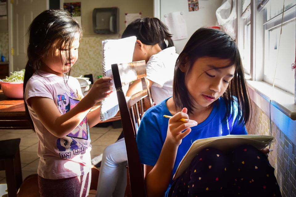 Eh Moo Taw Heh, right, draws in a notebook while her family prepares dinner in the kitchen on Oct. 25 at their home in Utica, N.Y. Eh Moo said her favorite class to take while learning from home was art.