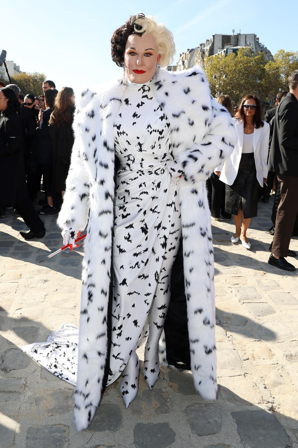 Alexis Stone dressed as Cruella de Vil in a spotted fur coat and long, flowing dress with similar pattern, holding a cigarette holder, posing outdoors at a likely public event