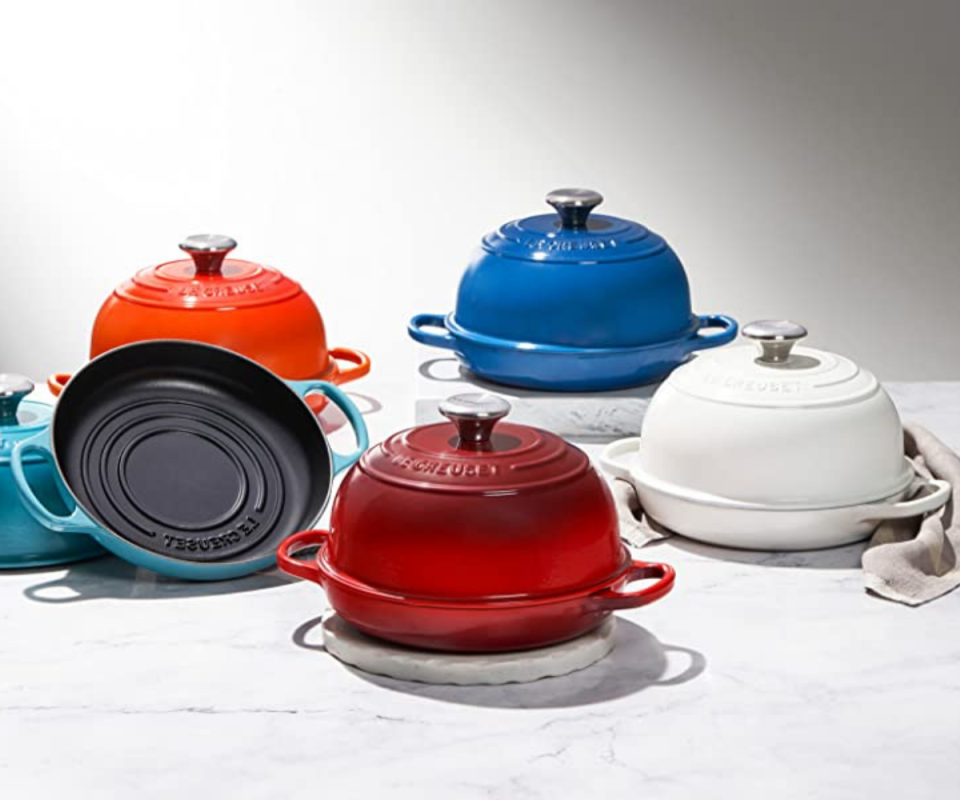 Four Le Creuset cast-iron bread ovens on a white marble table in blue, red, orange, navy and white with silver lids.