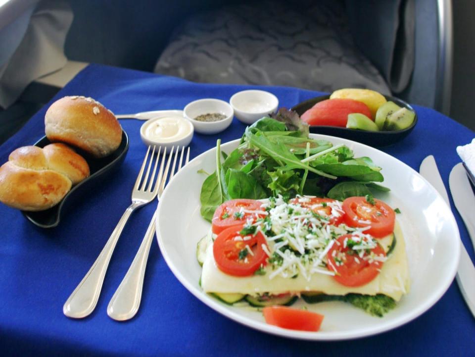 A stock image shows an in-flight meal, a pasta dish topped with tomatoes and served with greens, a plate of fruit, and a bread basket.