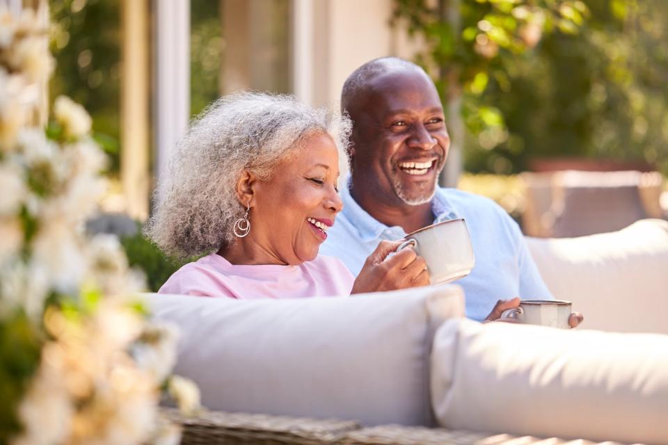 An elderly couple sits on a sofa outside and smiles.