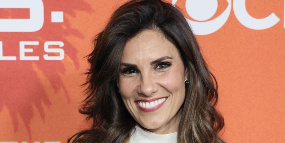 daniela ruah smiling at the camera in a white dress and camel coat