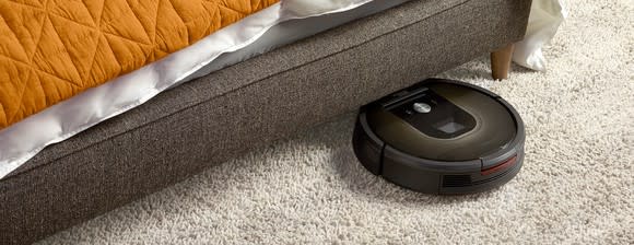 A Roomba robotic vac traveling on a rug and starting to go underneath an unidentifiable piece of furniture, possibly a bed.