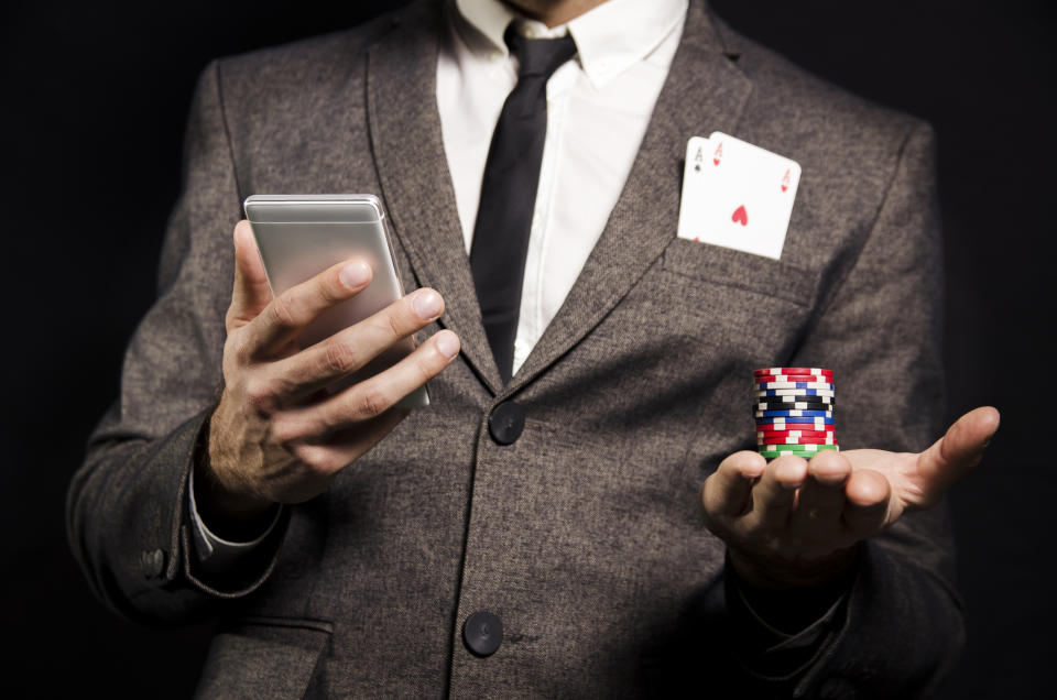 A man in a grey suit viewed from the neck down holds a smartphone in one hand and poker chips in the other and two aces in his jacket pocket.