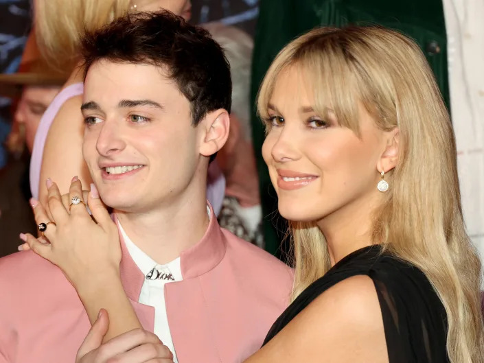 Noah smiling in a pink suit and Millie smiling in a black dress with her arm around Noah's chest.