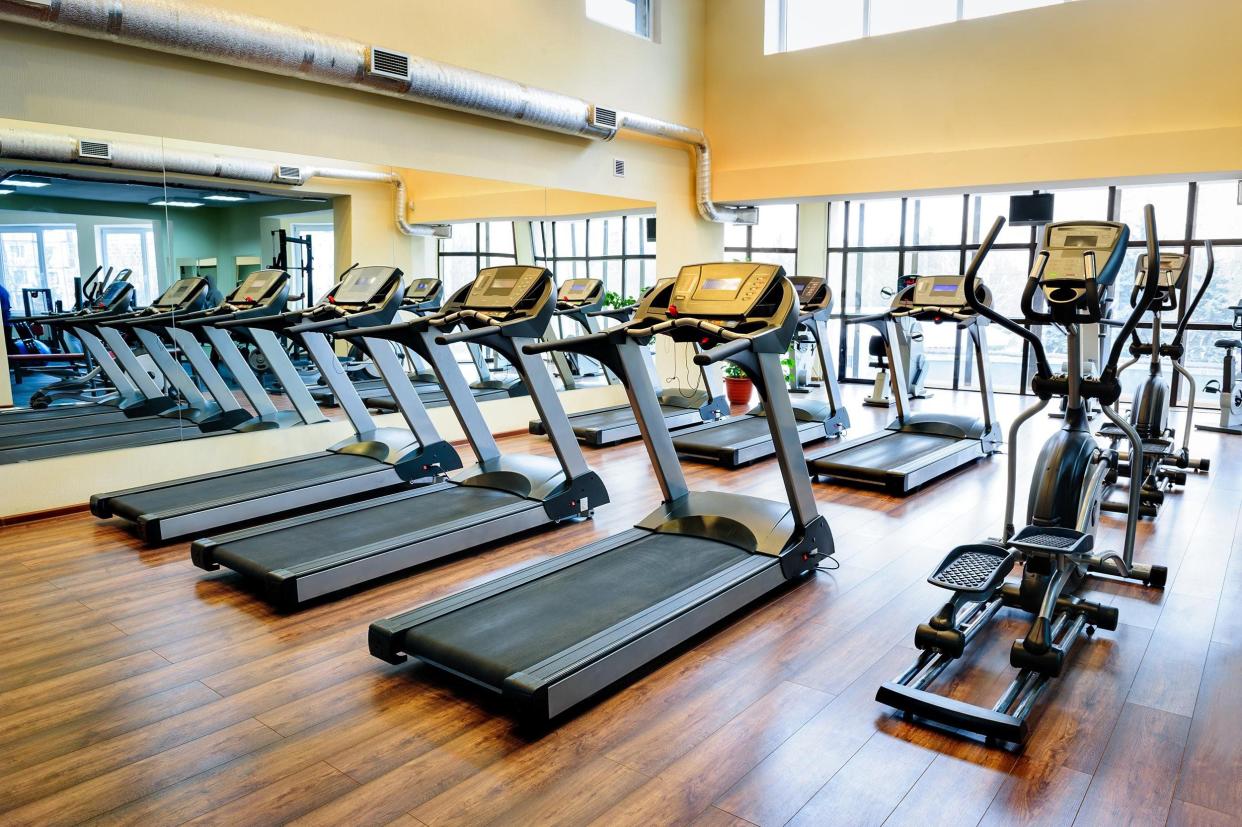 2 rows of treadmills with one elliptical each in an empty gym, facing a wall of windows, clean wooden floors and a high ceiling