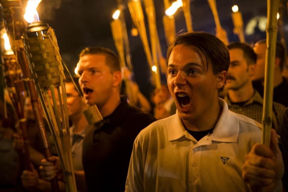 A group of people hold tiki torches during a nighttime rally, with one person in the foreground shouting passionately