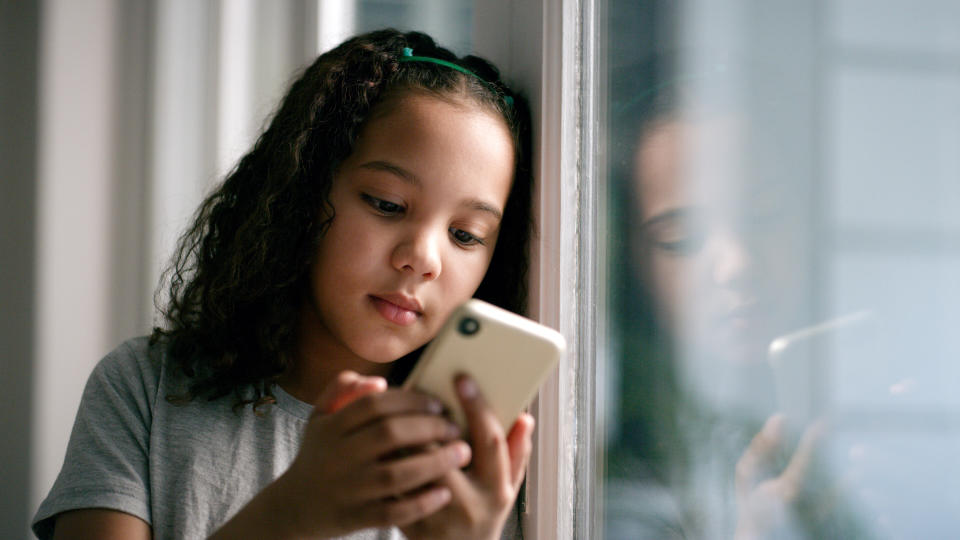 Window, smartphone and girl on social media, connection and search internet in house. Young female, kid and child with phone, online reading or communication being calm, playing games, device or chat