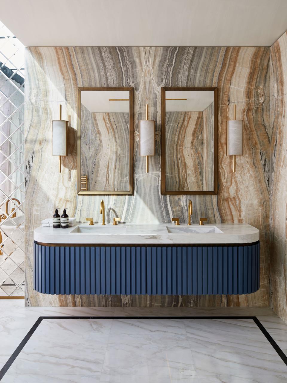 Custom notched wood vanity in French blue with marble and brass and sinks. Taps in polished brass are Volevatch. Sconces and mirrors by Humbert & Poyet; the walls are onyx.