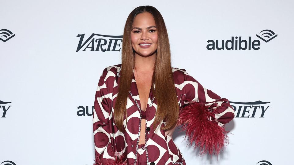 Chrissy Teigan in a red carpet photo for Variety and Audible. She's wearing a maroon and white geometric top with feathers on the sleeves. (Getty Images)