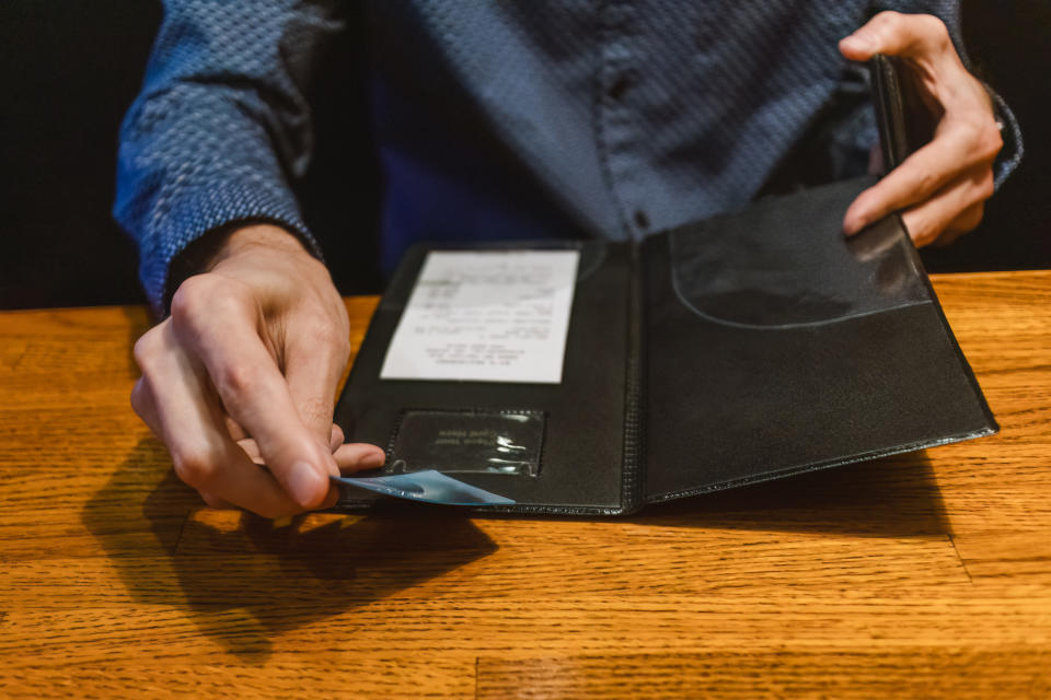 A person wearing a patterned shirt opens a restaurant bill booklet on a wooden table, revealing a receipt and payment card slot