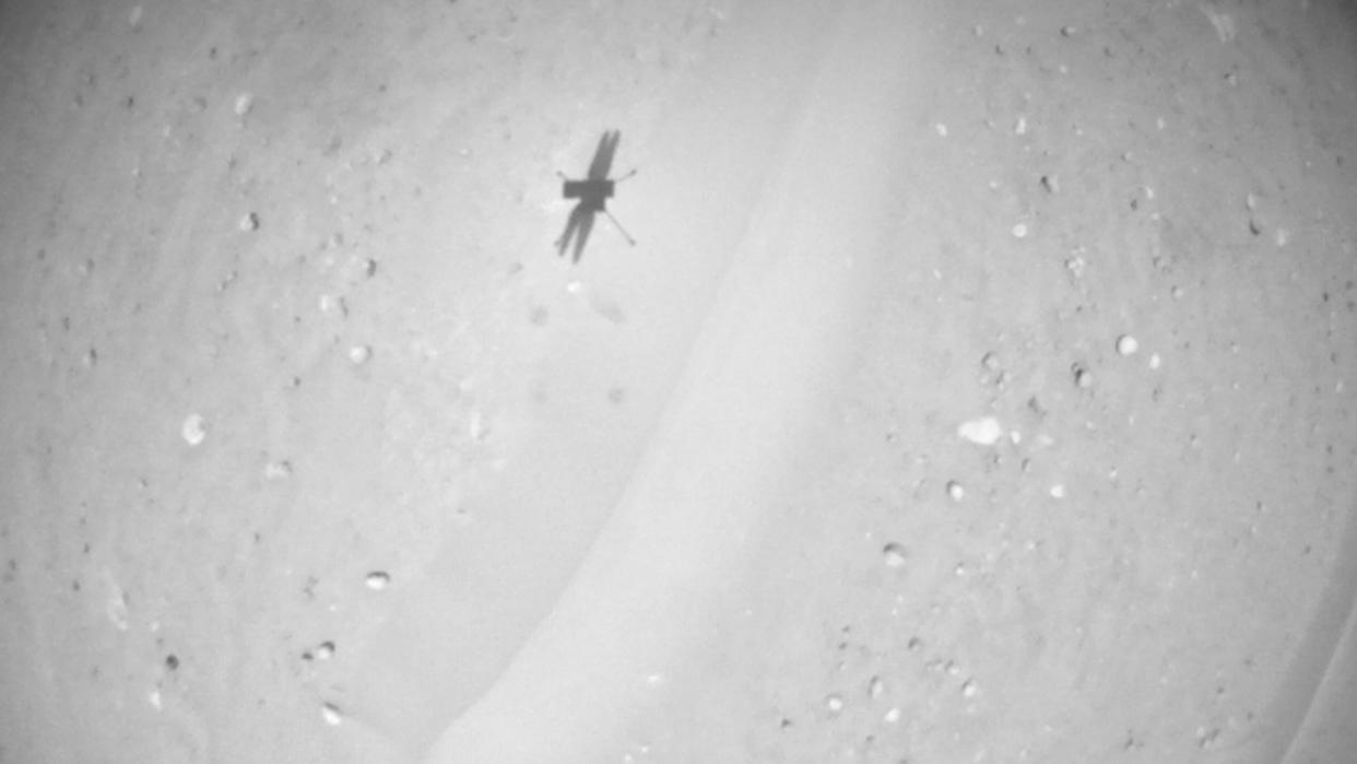  Black-and-white photo of a small drone's shadow on sandy, pebble-studded ground. 