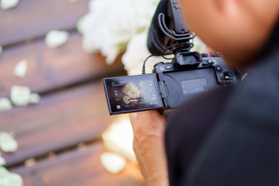 A photographer holds a camera, taking a close-up shot of wedding decorations or details, with the camera's screen showing the viewfinder image