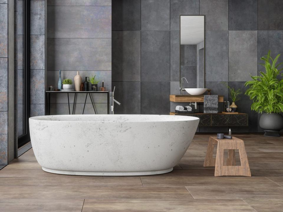 A bathroom with black and gray stone tiles on the walls, a wooden floor, and a light-gray stone bathtub